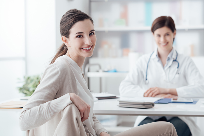 A woman sitting at a desk with a doctor in the background, discussing medical treatment.