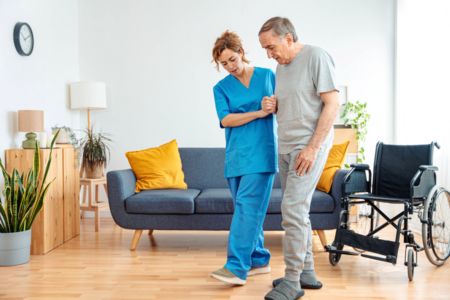 A female healthcare worker in scrubs assisting an elderly man in a wheelchair.