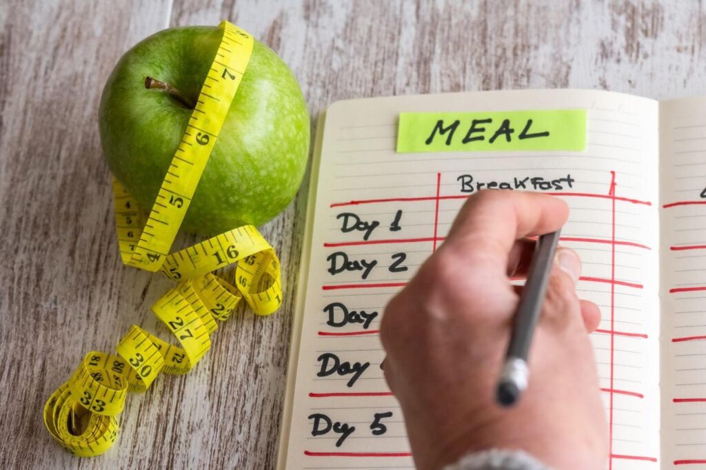 A hand writes a meal plan in a notebook next to an apple and measuring tape
