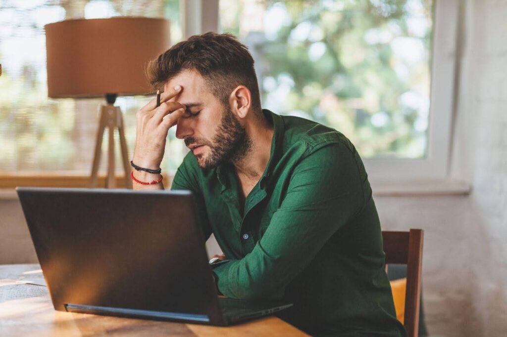 A man sits at a table head in hands looking stressed at his laptop